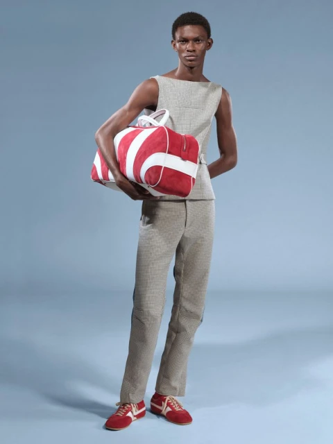 Adult male fashion model posing with a striped duffel bag. He is wearing a sleeveless top and pants, paired with matching red and white sneakers.