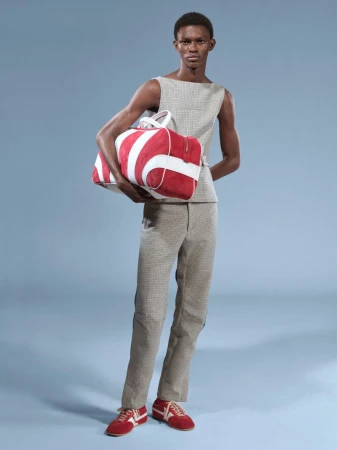 Adult male fashion model posing with a striped duffel bag. He is wearing a sleeveless top and pants, paired with matching red and white sneakers.
