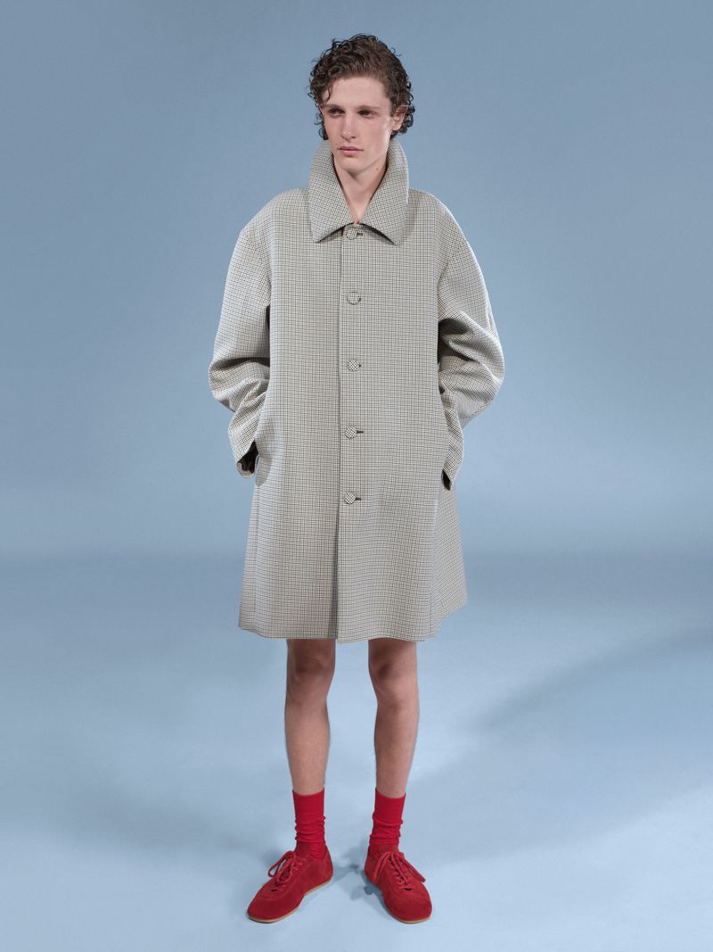 Man in a beige overcoat, red socks, and matching shoes, stands in a studio with a light blue backdrop. Fashion shoot, menswear.
