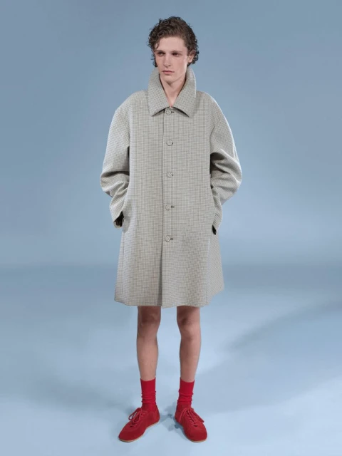 Man in a beige overcoat, red socks, and matching shoes, stands in a studio with a light blue backdrop. Fashion shoot, menswear.