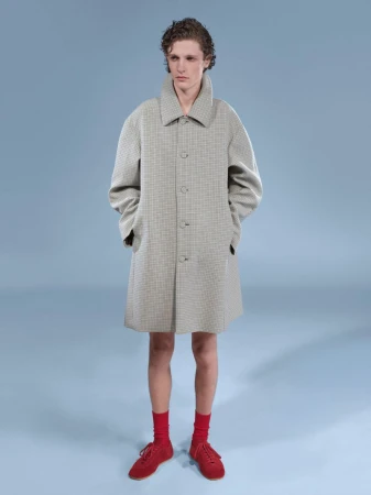 Man in a beige overcoat, red socks, and matching shoes, stands in a studio with a light blue backdrop. Fashion shoot, menswear.