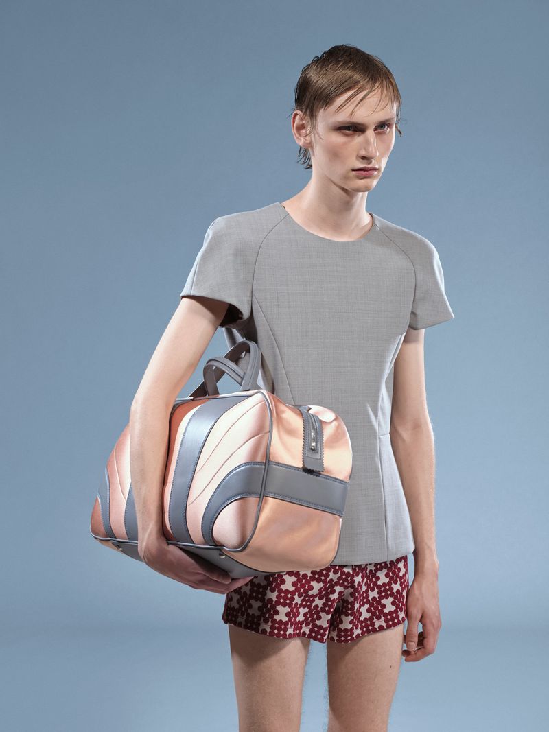 An adult male model poses in a grey shirt and patterned shorts, holding a pink-and-grey duffel bag against a plain blue backdrop in a studio shot.
