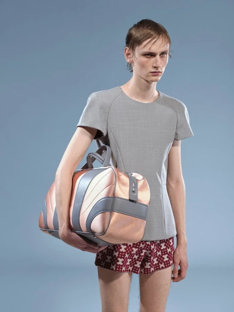 An adult male model poses in a grey shirt and patterned shorts, holding a pink-and-grey duffel bag against a plain blue backdrop in a studio shot.