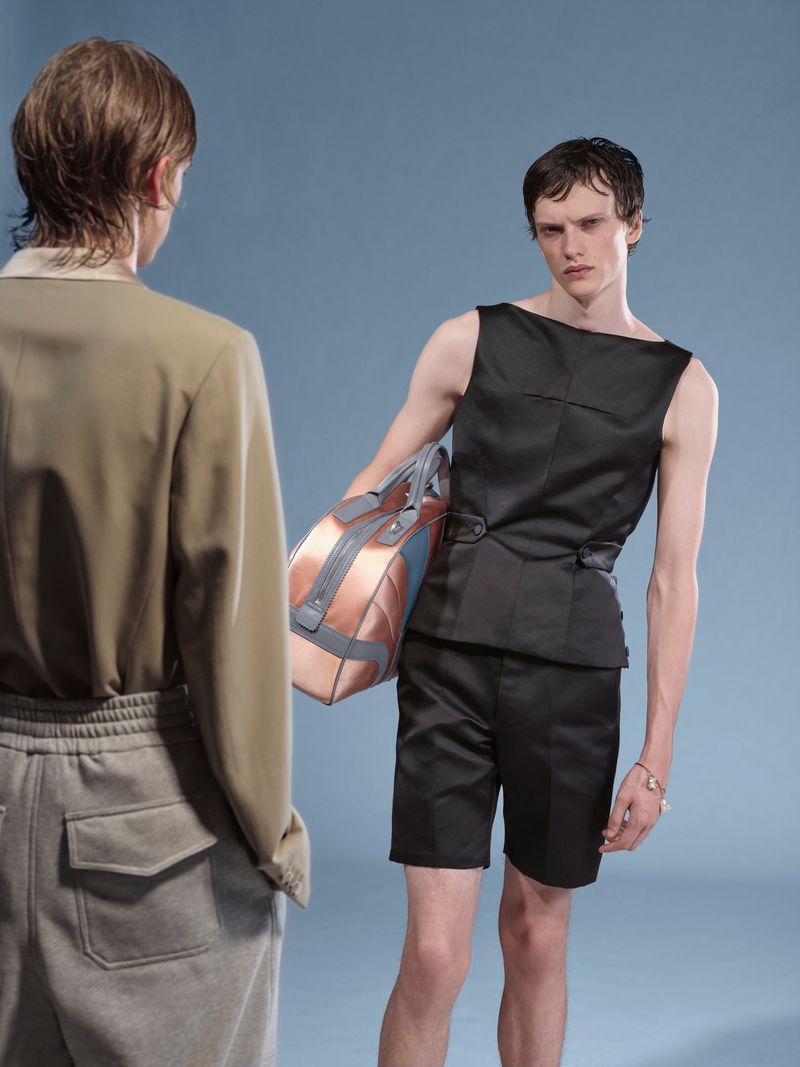 Two young men in contemporary fashion stand indoors against a light blue background in a medium studio shot with a model holding a bag.