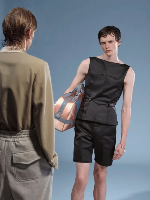 Two young men in contemporary fashion stand indoors against a light blue background in a medium studio shot with a model holding a bag.