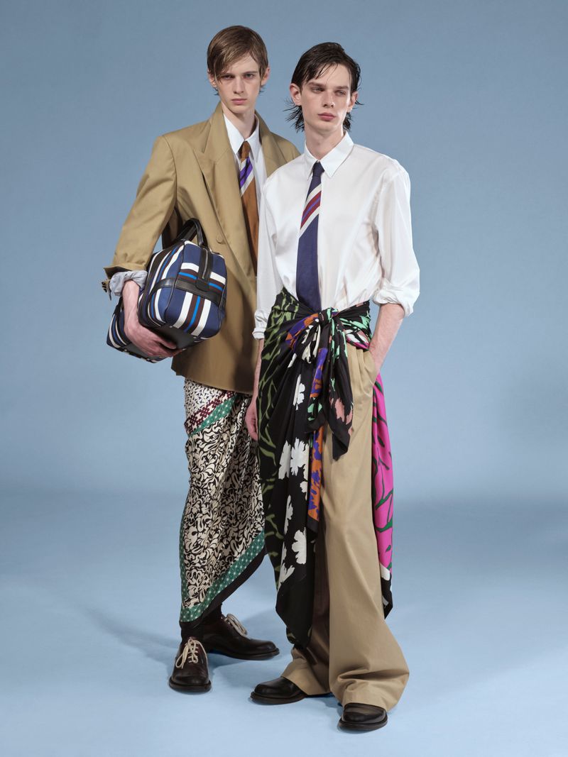 Two men pose in fashion shot with unique style, one in a blazer holding a bag, the other in a white shirt with a sarong. Light blue backdrop.