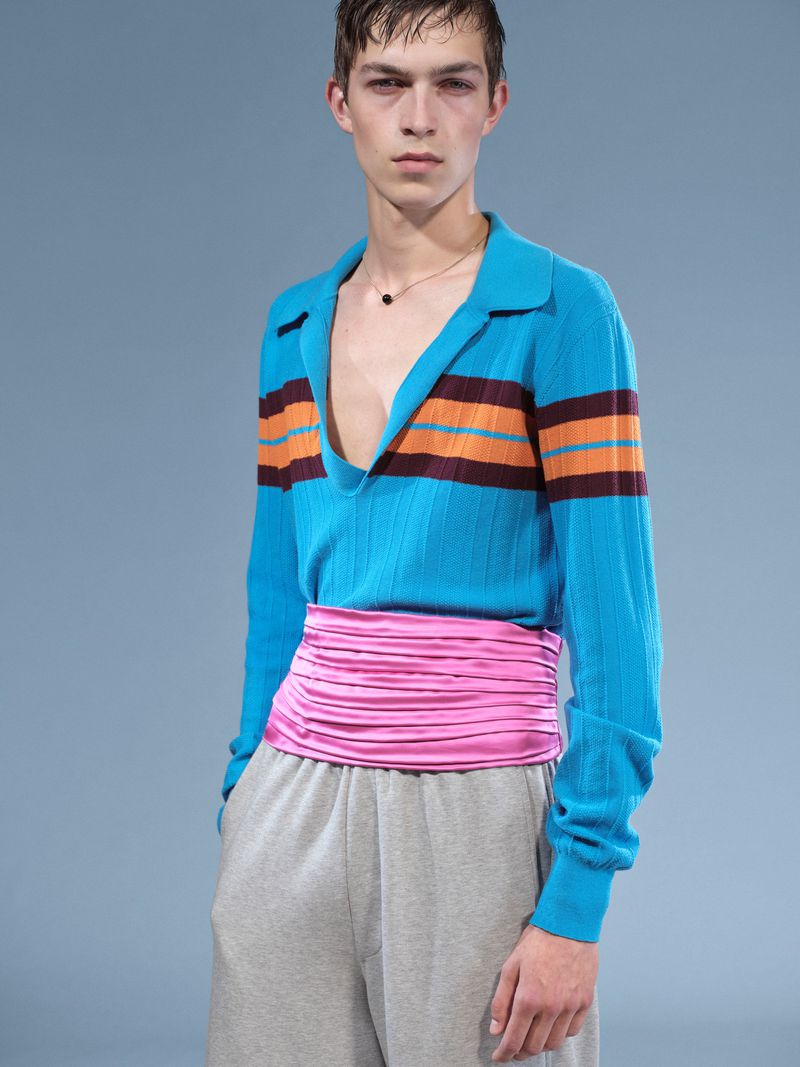 Young adult male fashion model poses in a turquoise striped polo shirt, pink waist wrap and gray sweatpants against a blue backdrop.