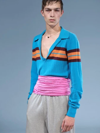 Young adult male fashion model poses in a turquoise striped polo shirt, pink waist wrap and gray sweatpants against a blue backdrop.