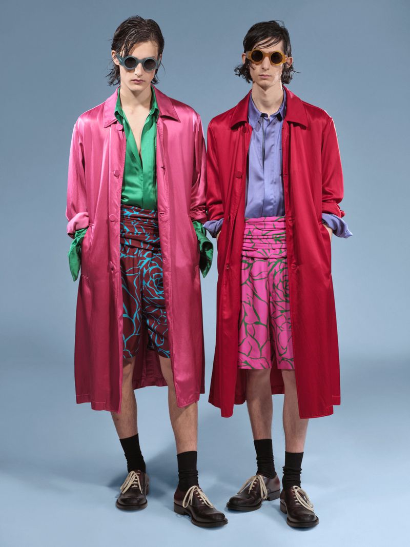 Two adult males stand side-by-side in a studio with a light-blue backdrop, both wearing sunglasses, overcoats, shorts, socks, and dress shoes.