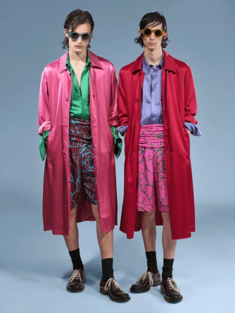 Two adult males stand side-by-side in a studio with a light-blue backdrop, both wearing sunglasses, overcoats, shorts, socks, and dress shoes.
