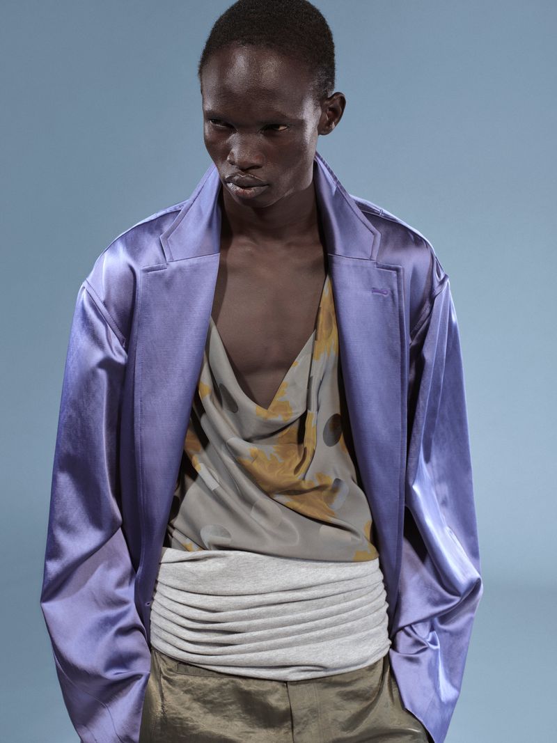 Fashion shot of a slender male model wearing a lavender jacket, top with a brown pattern, silver waist wrap, and olive pants against a blue backdrop.