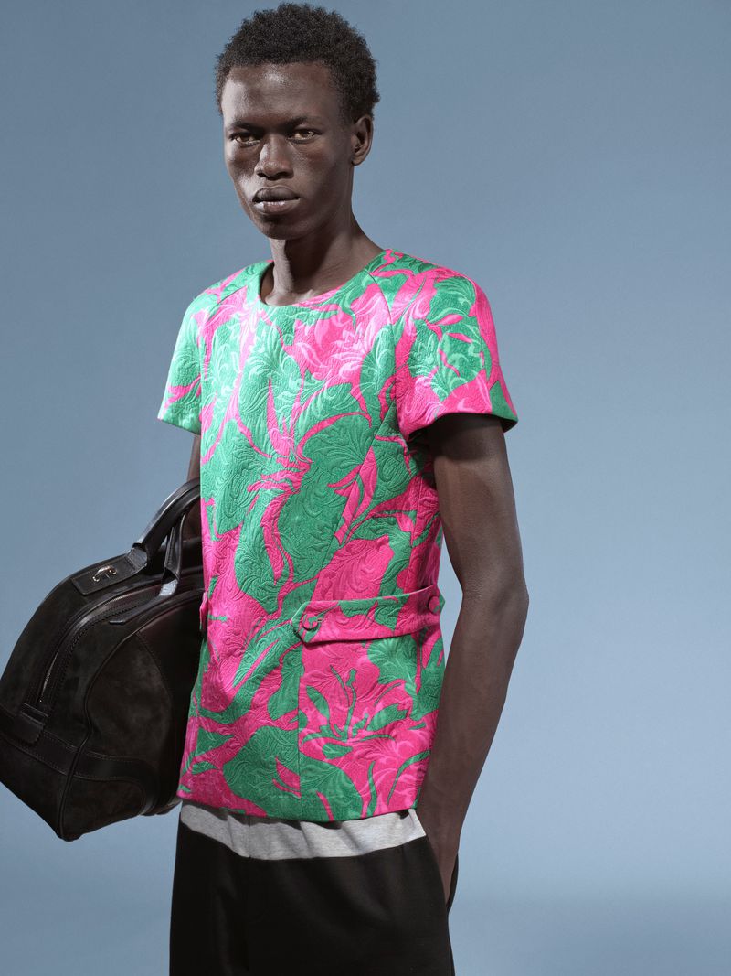 Studio shot of a dark-skinned male model in a pink and green shirt holding a black leather duffel bag against a blue background.