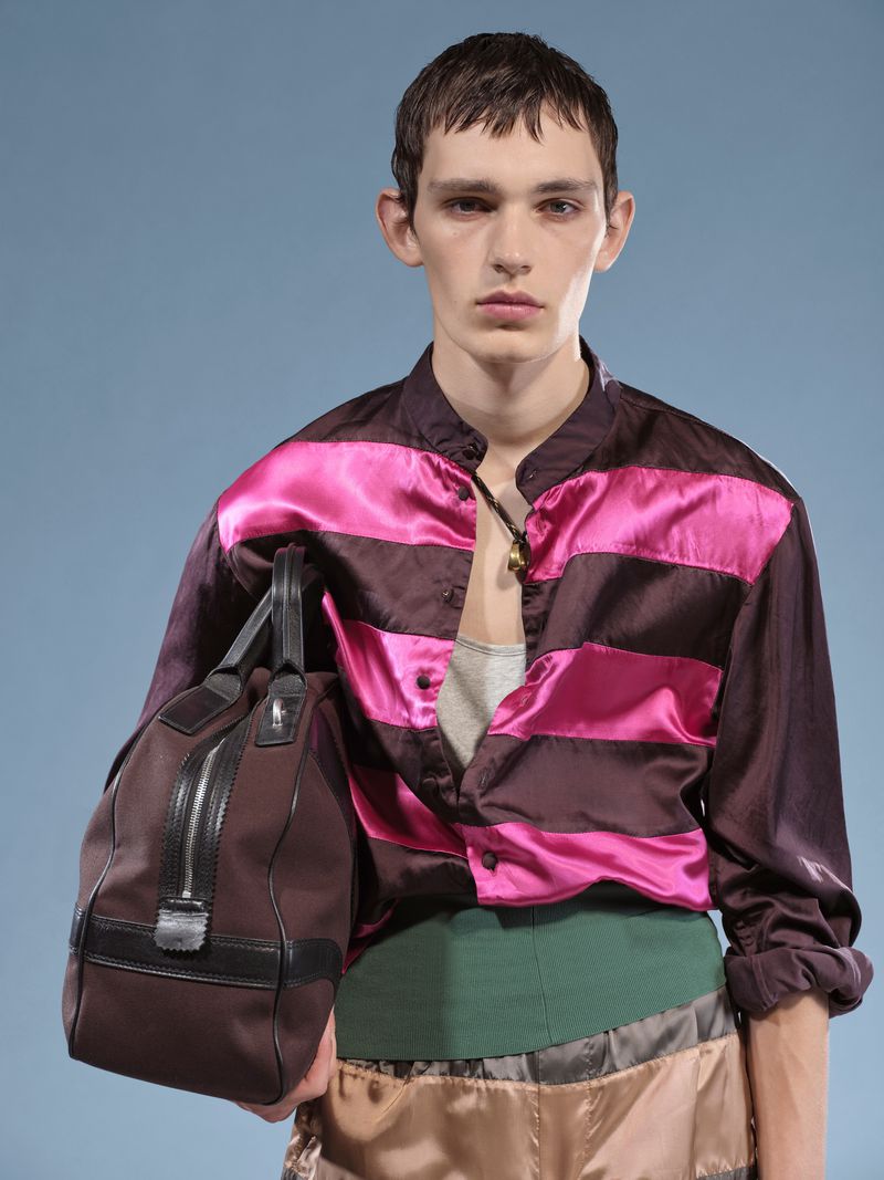 Young male fashion model posing with a brown duffel bag in a studio with a blue backdrop, wearing a shiny striped shirt.