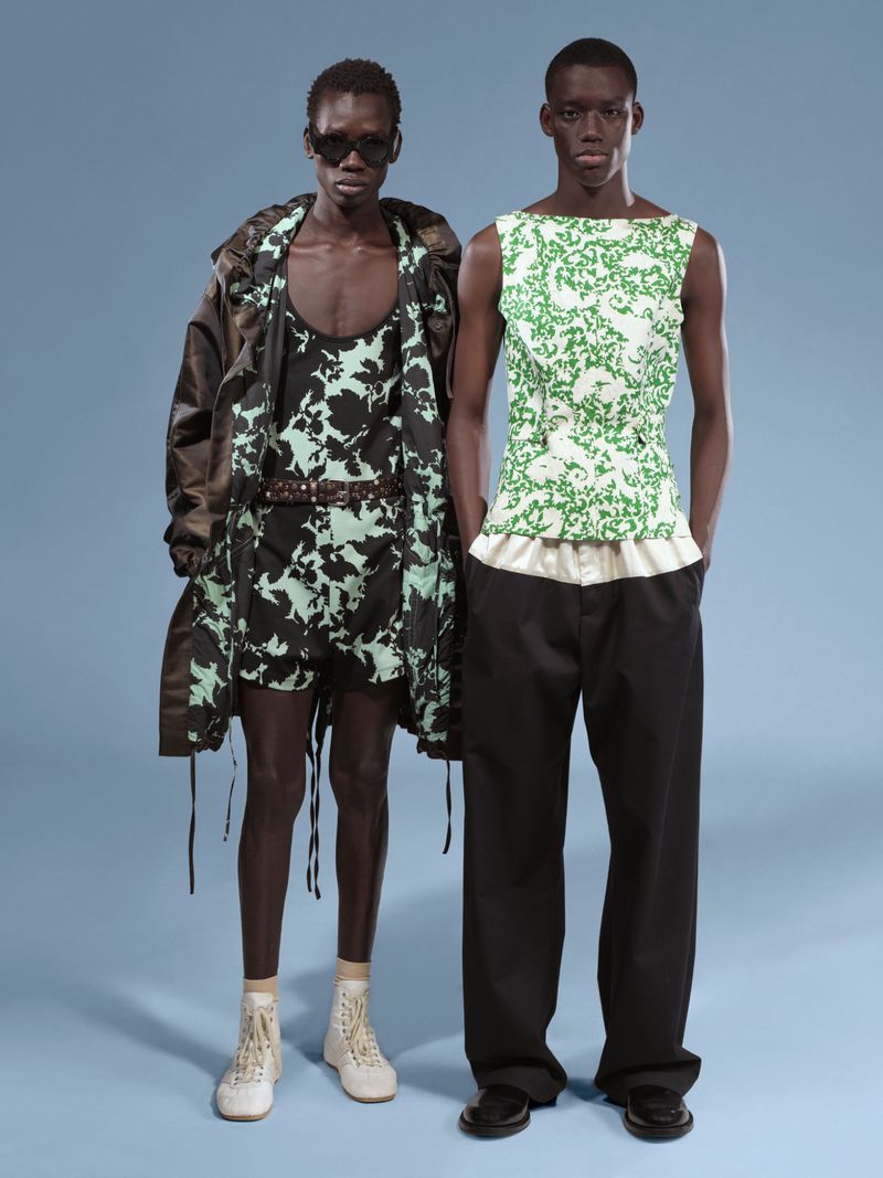 Two African male fashion models showcasing matching floral print tops and loose black pants against a light blue backdrop in a studio shot.