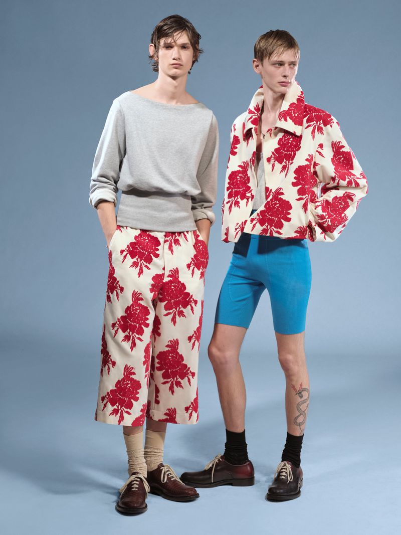 Two male fashion models pose in front of a blue background, wearing casual tops and patterned pants, with brown leather shoes.