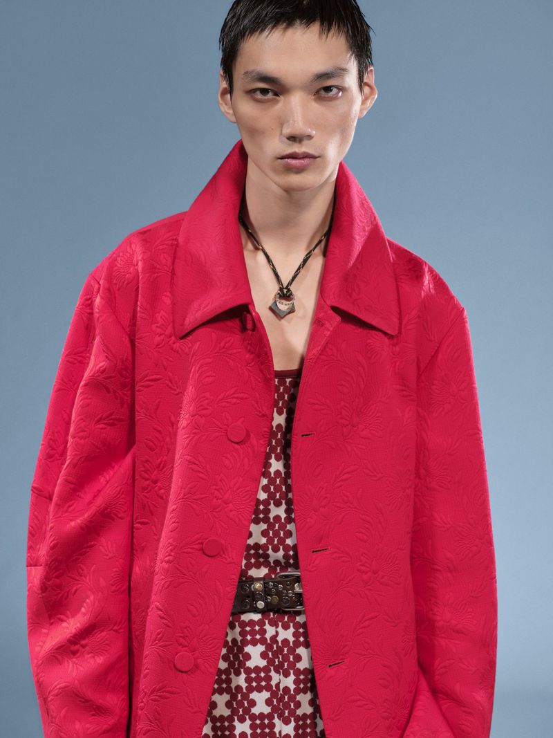 A single Asian male model in a bold red patterned coat and patterned shirt, standing in front of a light blue backdrop in a fashion shot.