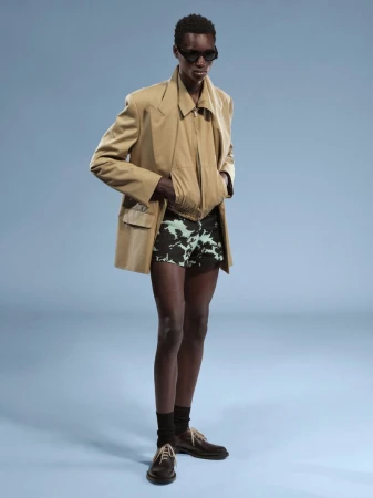 Model wearing a tan blazer, patterned shorts, sunglasses, and dark shoes against a light blue studio background.