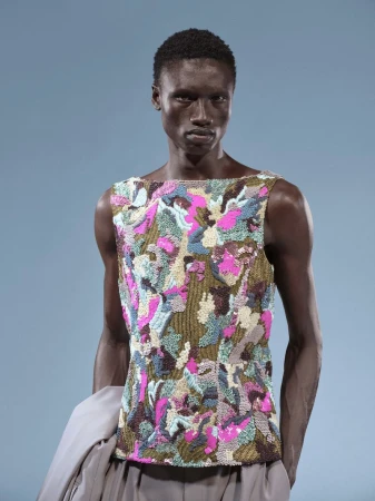 Model wearing a patterned multicolored sleeveless top and grey trousers against a solid blue background for Dries Van Noten lookbook