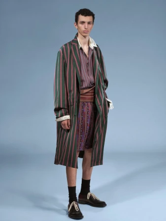 Model standing against a light blue backdrop wearing a striped green coat, patterned shorts, and dark shoes.
