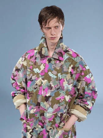 Model wearing a patterned textured coat with high collar against a plain neutral background