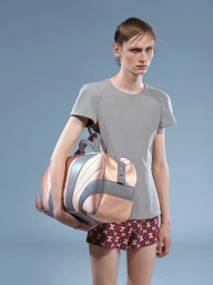 Male model in a grey short-sleeve top and patterned shorts carrying a large duffel bag against a light blue backdrop.