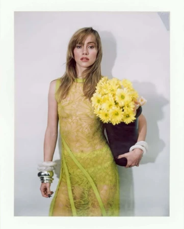 A slender woman with blonde hair poses in a green lace dress while holding a bouquet of yellow daisies in a black container.