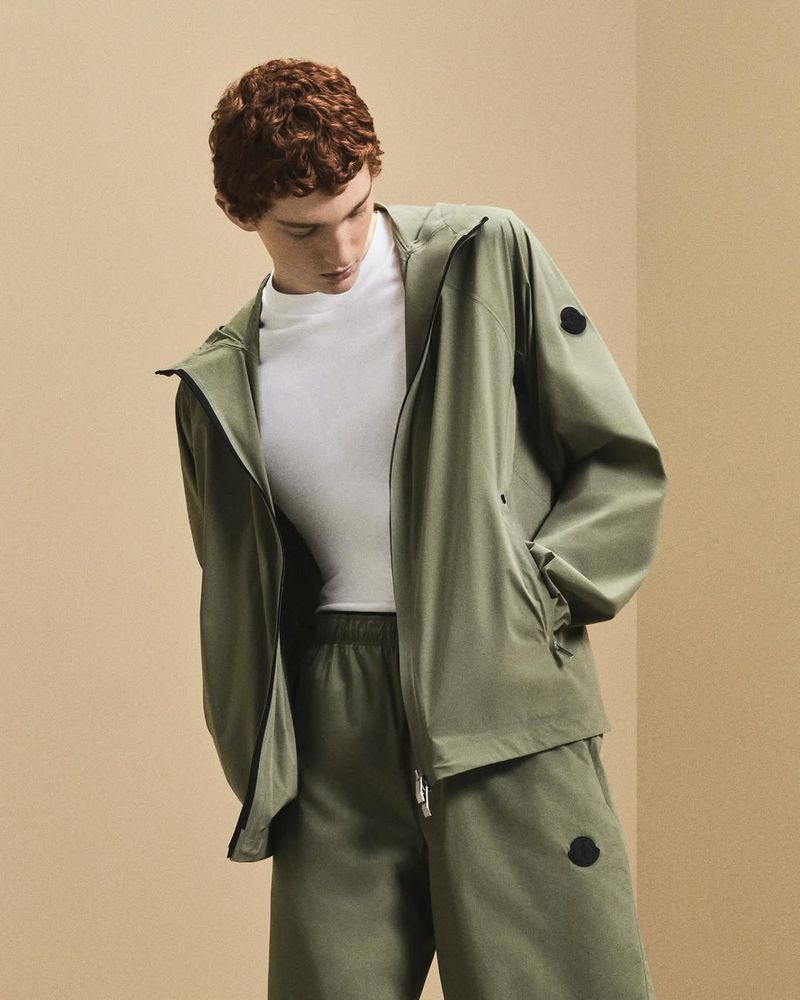 A slender male model in a light-olive tracksuit with a matching zip-up jacket stands indoors against a light-tan wall for a fashion advertisement.