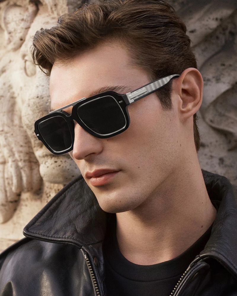 Close-up of a fashionable adult man with sunglasses. He is wearing a black leather jacket, standing against an ornate background.