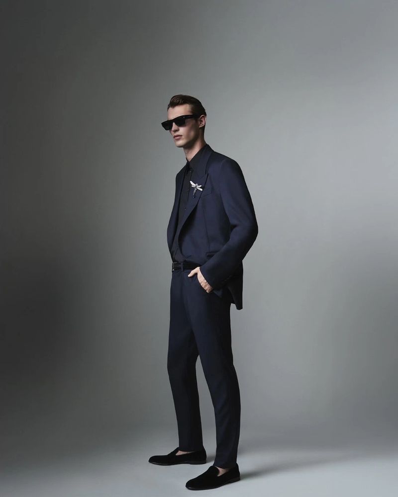 A male model in a dark suit and sunglasses poses in a studio. He is looking to the side with one hand in his pocket.