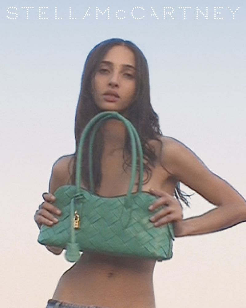 A slender woman poses with a green woven handbag against a pale blue sky in a fashion editorial campaign, featuring the Stella McCartney logo.