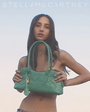 A slender woman poses with a green woven handbag against a pale blue sky in a fashion editorial campaign, featuring the Stella McCartney logo.