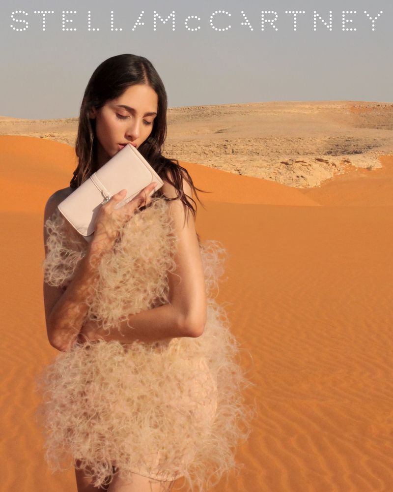 Fashion shot of woman holding a purse, posing in a dress made of feathers, with a desert landscape in the background, under daylight.
