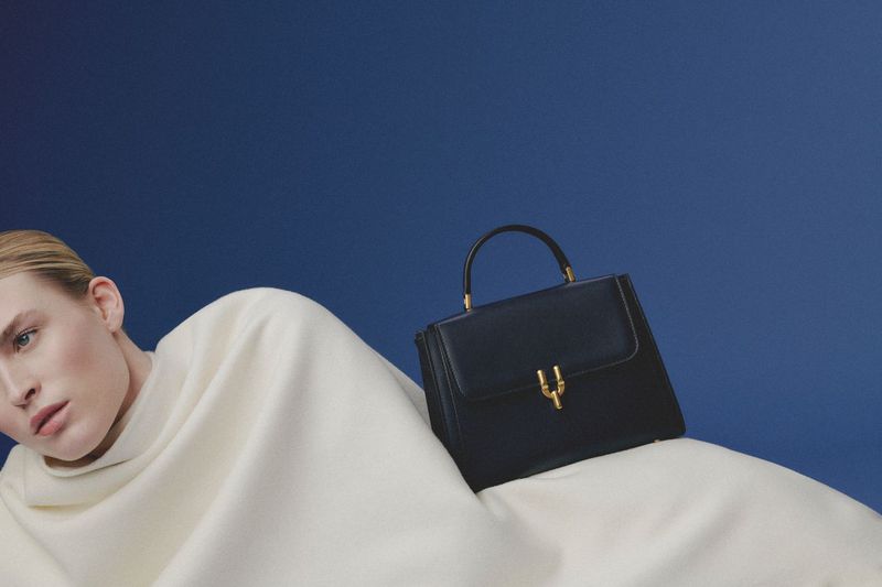 Fashion shot of a blonde woman lying down next to a black leather handbag with gold hardware against a dark blue background.