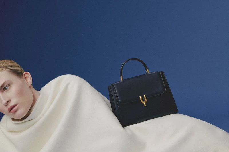 Fashion shot of a blonde woman lying down next to a black leather handbag with gold hardware against a dark blue background.