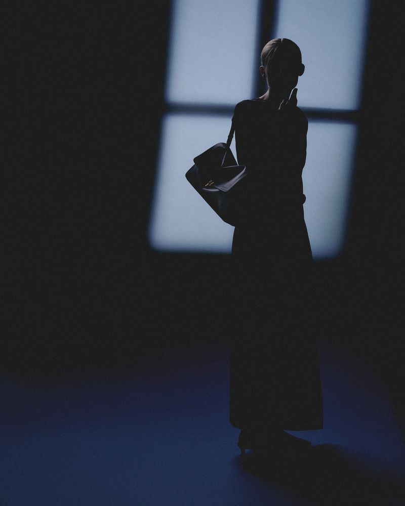 Silhouette of an adult female standing with a purse in front of a window casting light in an otherwise dark studio setting.