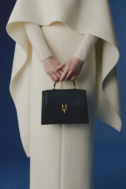 Elegant fashion photo showcasing a woman in a white dress and cape holding a structured black handbag against a gradient blue backdrop.