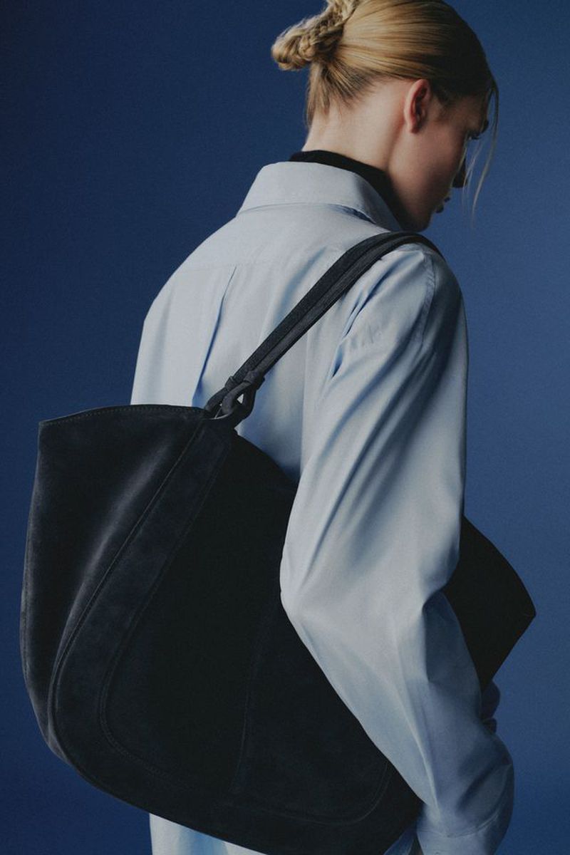 Model wearing a light blue shirt carrying a large dark suede shoulder bag against a deep blue background.