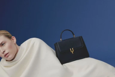 Close-up of a blonde model partially draped in cream fabric next to a dark handbag with gold hardware against a blue background.