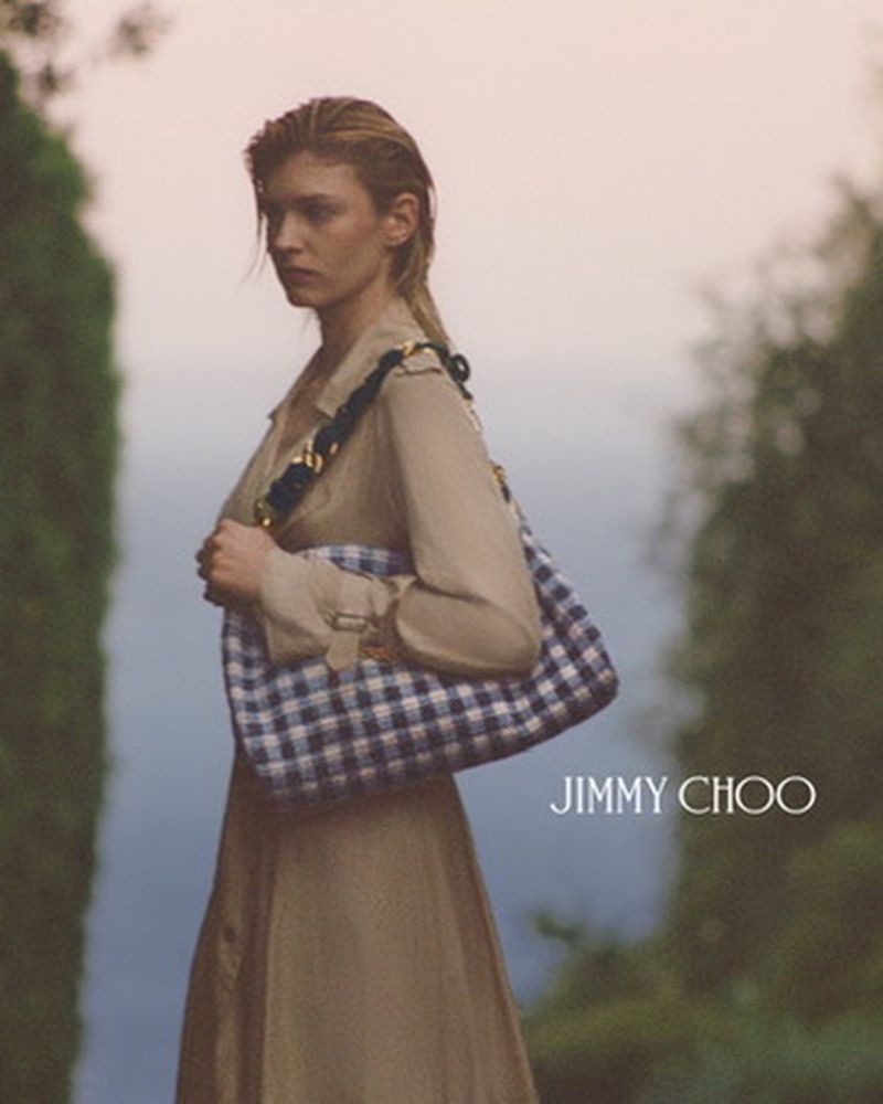Woman wearing a tan trench coat and a blue and white gingham tote bag outdoors with the Jimmy Choo logo visible on the lower right.