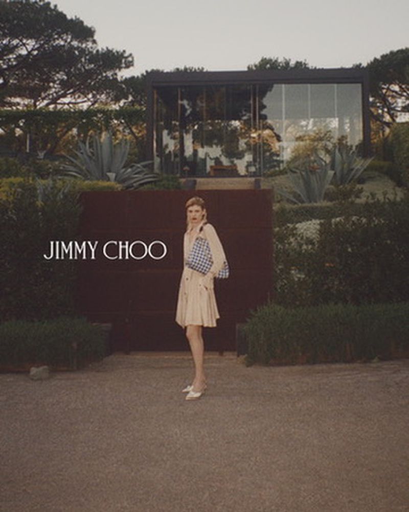 Elegant woman in a trench coat and handbag stands in front of a modern house surrounded by green foliage. Jimmy Choo branding.