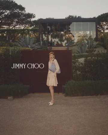Elegant woman in a trench coat and handbag stands in front of a modern house surrounded by green foliage. Jimmy Choo branding.