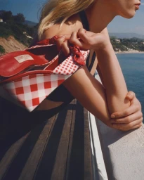 Model holding a red and white gingham wedge shoe while leaning against a balcony overlooking the ocean.