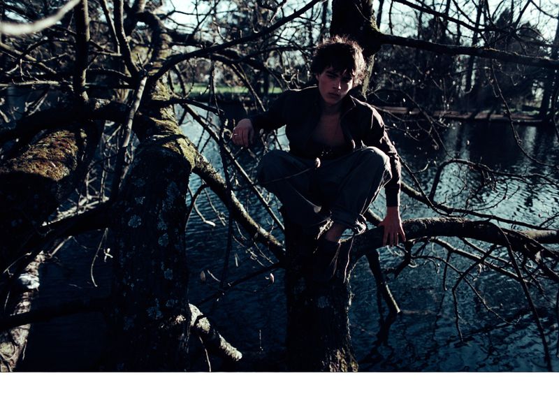 Man sits in a crouched position on a tree branch in an outdoor setting, with a body of water visible in the background.