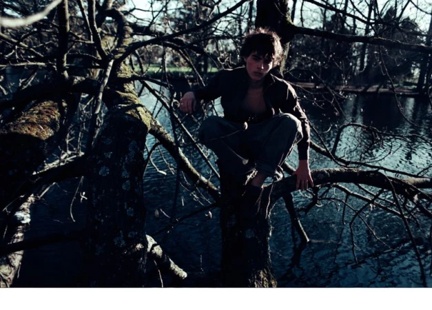 Man sits in a crouched position on a tree branch in an outdoor setting, with a body of water visible in the background.