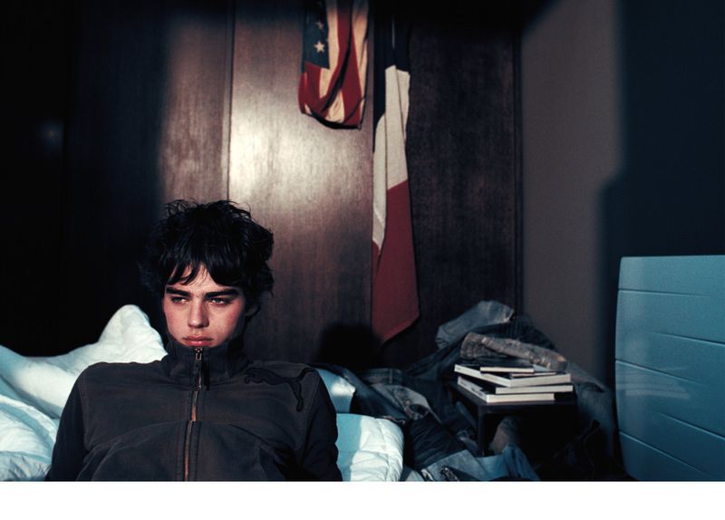 Young man in a jacket on a bed indoors, American and French flags hanging on the wall, creating an intimate, somewhat somber atmosphere.