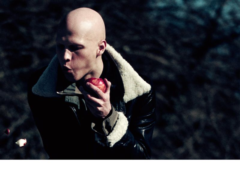 A bald person in a dark coat holds a red apple outdoors with a dark, blurred background. Some red and white objects float in foreground.