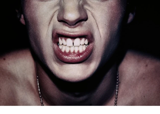 Close-up of an adult with gritted teeth and exposed gums, wearing a beaded necklace against a stark black background.