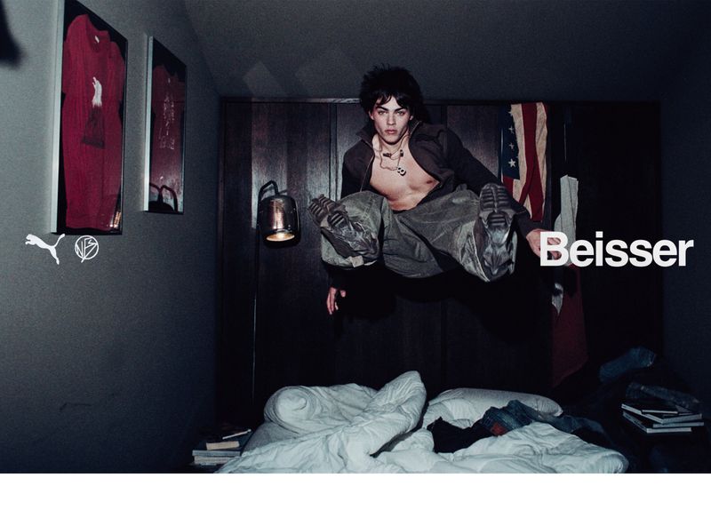 Man in mid-air indoors wearing neutral clothing, bed below, posters on wall, American flag in background, brand name 'Beisser'.
