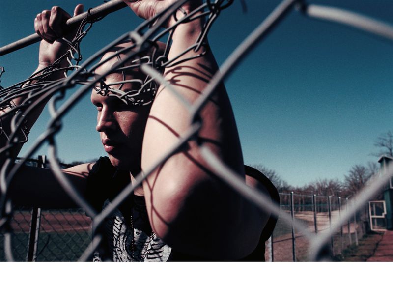 Low-angle shot of a young adult male holding a metal fence with blue sky background and green trees in the distance, filtered.