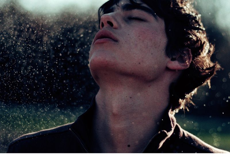 Close-up portrait of a young adult male with wet hair and closed eyes, as water droplets fall on his face, outdoors in soft lighting.